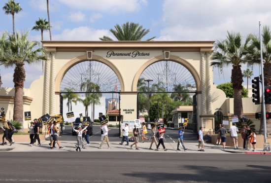 A line of protestors holding picket signs march in front of the entrance to the paramount pictures studio